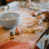 Nahaufnahme eines mit Mehl bestäubten Holztischs, auf dem eine Keramikschale, ein kleiner Nudelholz sowie zwei mehlbedeckte Holzakornfiguren des Produkts Freies Spiel: Bakes von KLICKKLACK neben einer Kinderhand liegen.