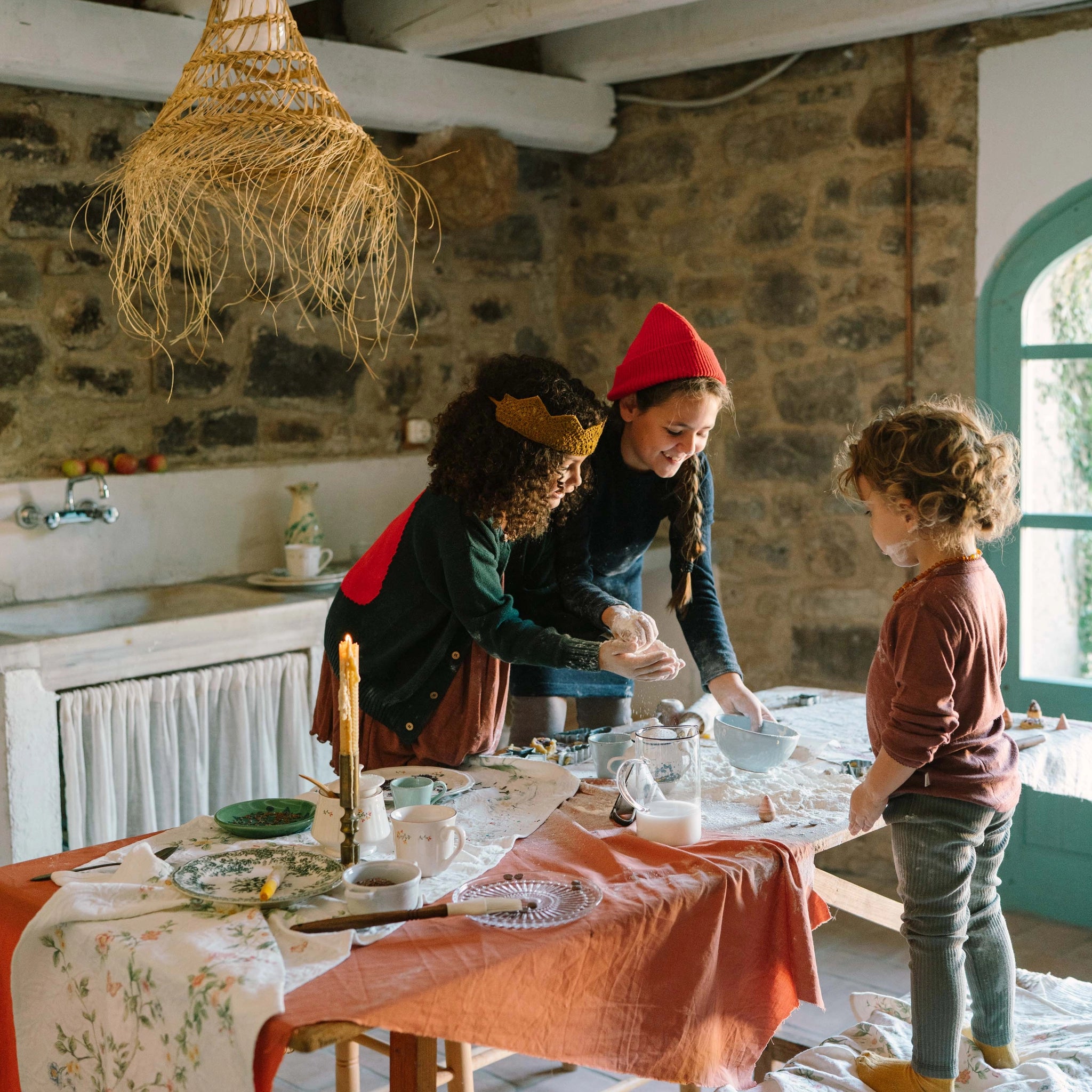 Drei Kinder backen fröhlich am rustikalen Tisch mit dem Set Freies Spiel: Bakes, das Schüsseln, Nudelholz und Ausstechformen umfasst, während sie Teig kneten. Ideal für kreative Backerlebnisse.