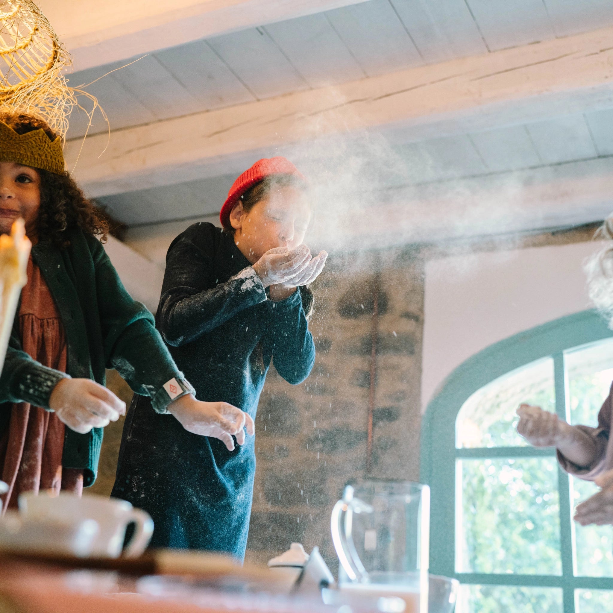 Kinder genießen Freies Spiel: Bakes, während ein Mädchen in roter Mütze Mehl in die Luft pustet. Der Tisch ist mit Backutensilien bedeckt, was eine spielerische, mehlverschmierte Atmosphäre schafft.