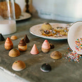 Freies Spiel: Bakes - Auf einer Arbeitsplatte liegen hölzerne Stapel-Eiswaffeln mit pastellfarbenen Kugeln. Daneben ein umgestürztes Schälchen mit Streuseln und ein dekorativer Teller mit Gebäck im Hintergrund.