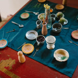 Freies Spiel: Dishes – Eine rustikale Anordnung von bemalten Holztellern, winzigen Löffeln, Metall- und Keramikgefäßen sowie natürlichen Akzenten auf einem Tisch, verkörpert nachhaltiges Design und zeitlose Ästhetik.