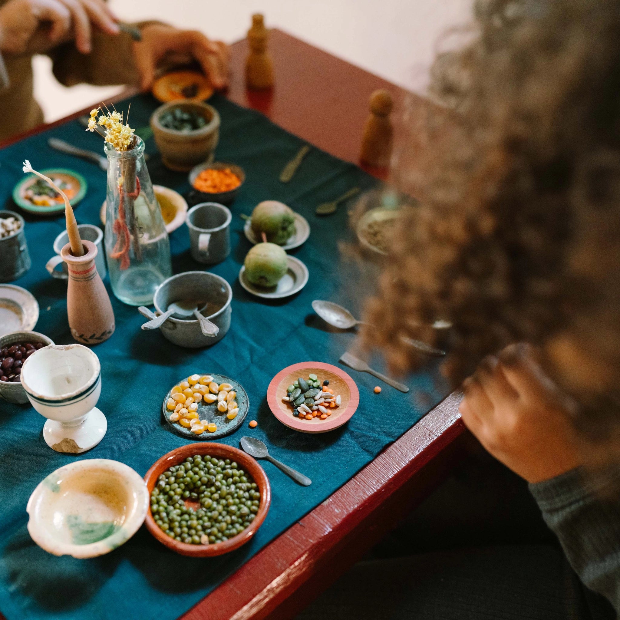 Freies Spiel: Dishes präsentiert eine Sammlung kleiner Schalen und Teller, die grüne Erbsen, gelbe Maiskörner, Samen und Linsen enthalten, begleitet von Perlen, Löffeln und einem Kerzenhalter.