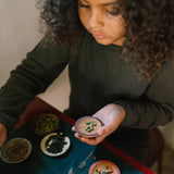 Ein Kind sitzt an einem kleinen Tisch und spielt mit Freies Spiel: Dishes. Vor ihr liegen Schalen mit verschiedenen Hülsenfrüchten und Samen, die sie mit einem hölzernen Spatel sortiert.