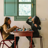 Zwei Kinder spielen an einem rustikalen roten Tisch mit Freies Spiel: Dishes, einer Geschirrkollektion. Eine Vase mit Trockenblumen und kleine Tassen und Schalen gestalten eine fantasievolle Teeparty-Atmosphäre.