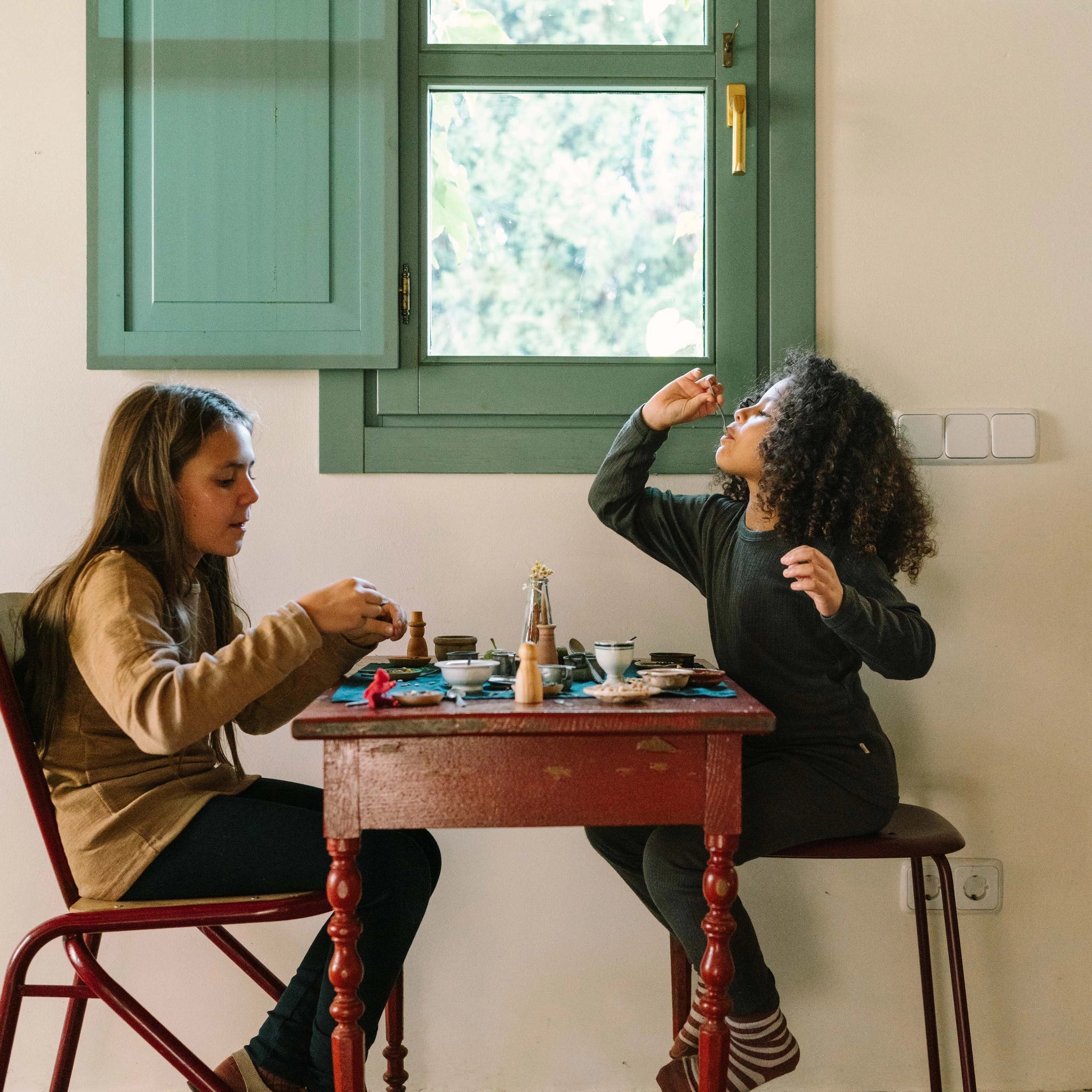 Zwei Kinder spielen an einem rustikalen roten Tisch mit Freies Spiel: Dishes, einer Geschirrkollektion. Eine Vase mit Trockenblumen und kleine Tassen und Schalen gestalten eine fantasievolle Teeparty-Atmosphäre.