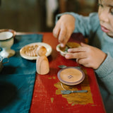 Kinderspiel-Setup auf abgenutztem roten Holztisch: Freies Spiel: Dishes mit Holzfigur, kleinem Teller, Mini-Besteck und Schale mit grünen Spiel-Lebensmitteln, daneben Eierbecher und Schälchen mit Müsli oder Nüssen.