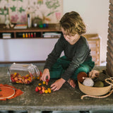 Ein Kind spielt mit dem Freies Spiel: Warm Mandala Mix, sortiert und stapelt spindelförmige Holzstücke auf einer niedrigen Steinbank. Nebenbei liegen eine Stickerei und ein Korb mit Garn.