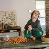 Ein fröhliches Kind sitzt auf einem Tisch und fädelt die Freies Spiel: Warm Mandala Mix-Holzteile auf eine Schnur. Die warmen Holzteile sind aus einem umgekippten Glasbehälter verstreut.