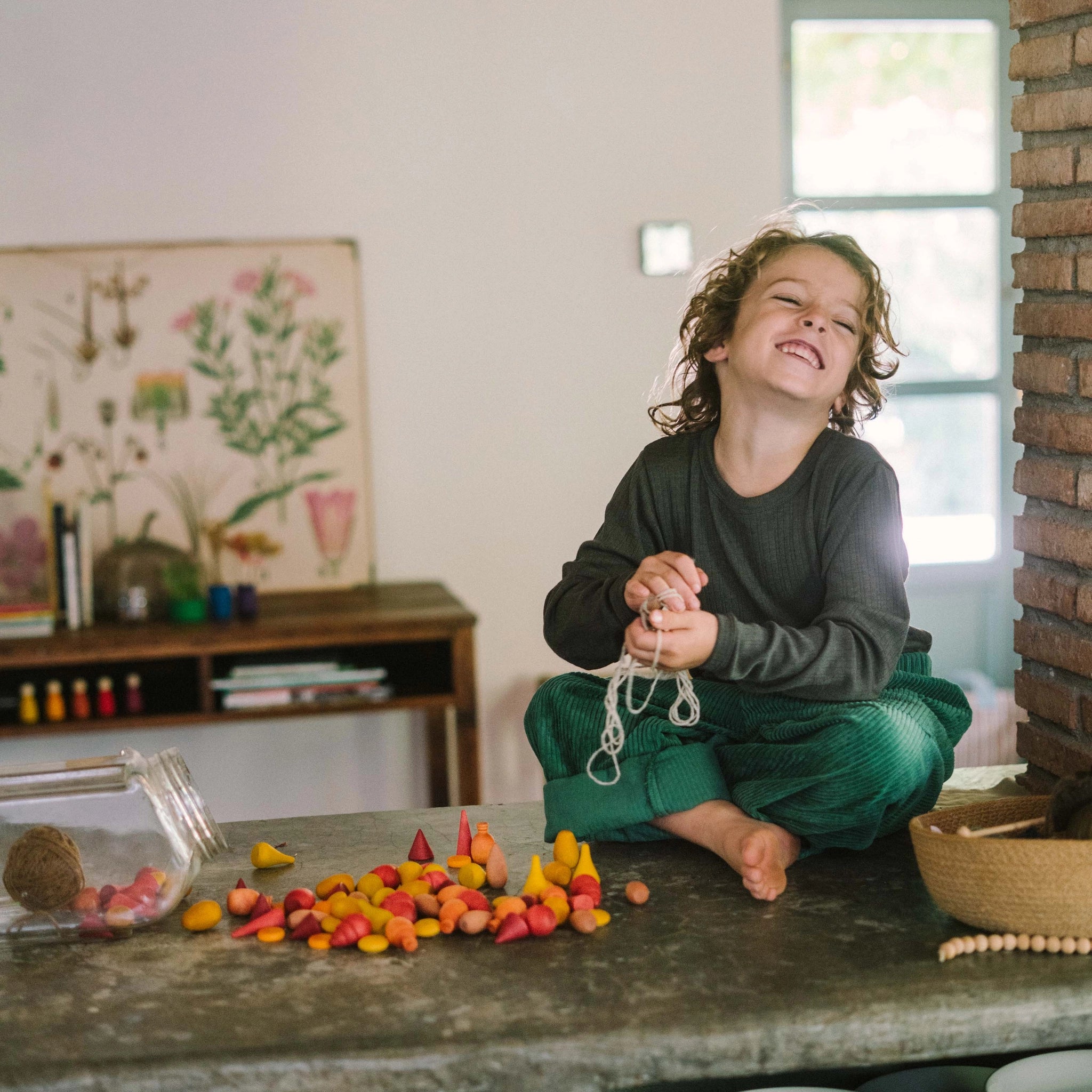 Ein fröhliches Kind sitzt auf einem Tisch und fädelt die Freies Spiel: Warm Mandala Mix-Holzteile auf eine Schnur. Die warmen Holzteile sind aus einem umgekippten Glasbehälter verstreut.