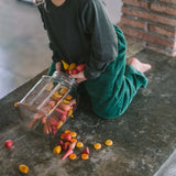 Kind kniet auf Beton, sammelt warmfarbene Freies Spiel: Warm Mandala Mix Holz- oder Wachsstifte, die aus einem umgekippten Glasgefäß gefallen sind, in einem grünen Cordkleid von KLICKKLACK.