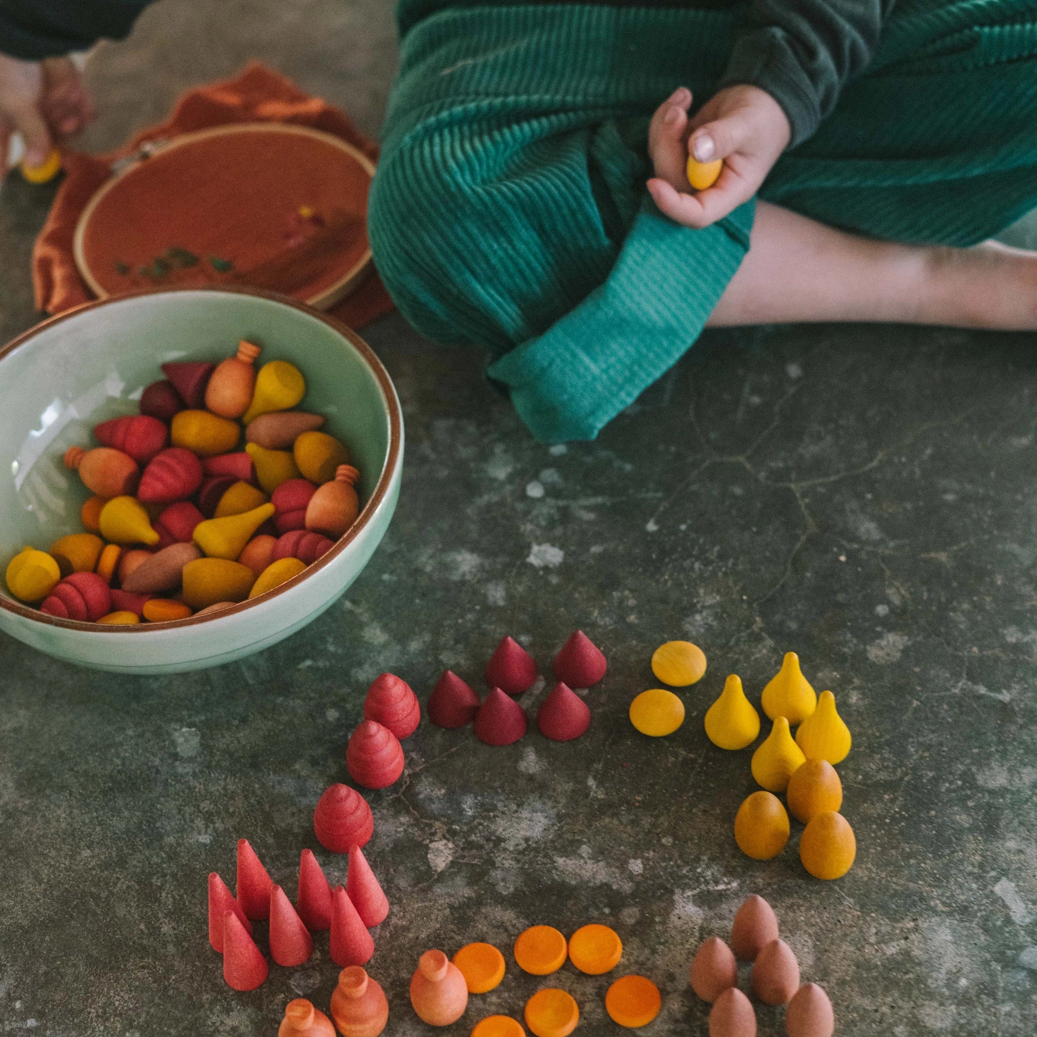 Ein Kind sortiert auf dem Boden mit Freies Spiel: Warm Mandala Mix aus Holz. Die Spielfiguren in Tropfen-, Kegel- und Scheibenform werden zu farblichen Gruppen arrangiert.
