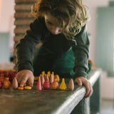 Ein Kind ordnet die leuchtend bunten Holzformen des Freies Spiel: Cold Mandala Mix auf einem Tisch. Die Formen umfassen Kegel, Tropfen und Spindeln, die kreative Spielfreude fördern.