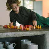 Kind liegt auf Betontheke und arrangiert Freies Spiel: Cold Mandala Mix, eine Sammlung kleiner, hölzerner Formen. Unter der Theke stehen gestapelte weiße Keramikschalen und passende Tassen.