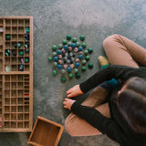 Ein Kind arrangiert bunte, runde Holzstücke in konzentrischen Kreisen auf dem Boden. Die „Freies Spiel: Moonlight“-Teile liegen neben einer Holzschale mit Fächern auf dem Boden.