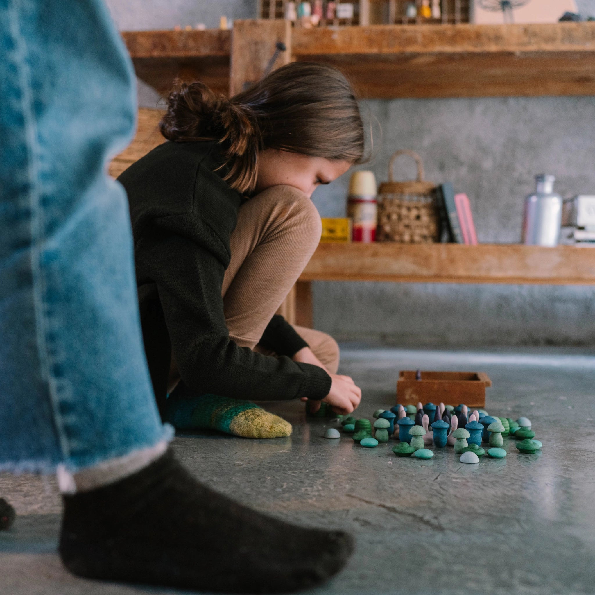 Ein Kind ordnet sorgfältig Freies Spiel: Moonlight, bestehend aus pilz- und kieselsteinförmigen Teilen, auf dem Boden. Im Hintergrund sind Regale mit Körben und Büchern sichtbar.