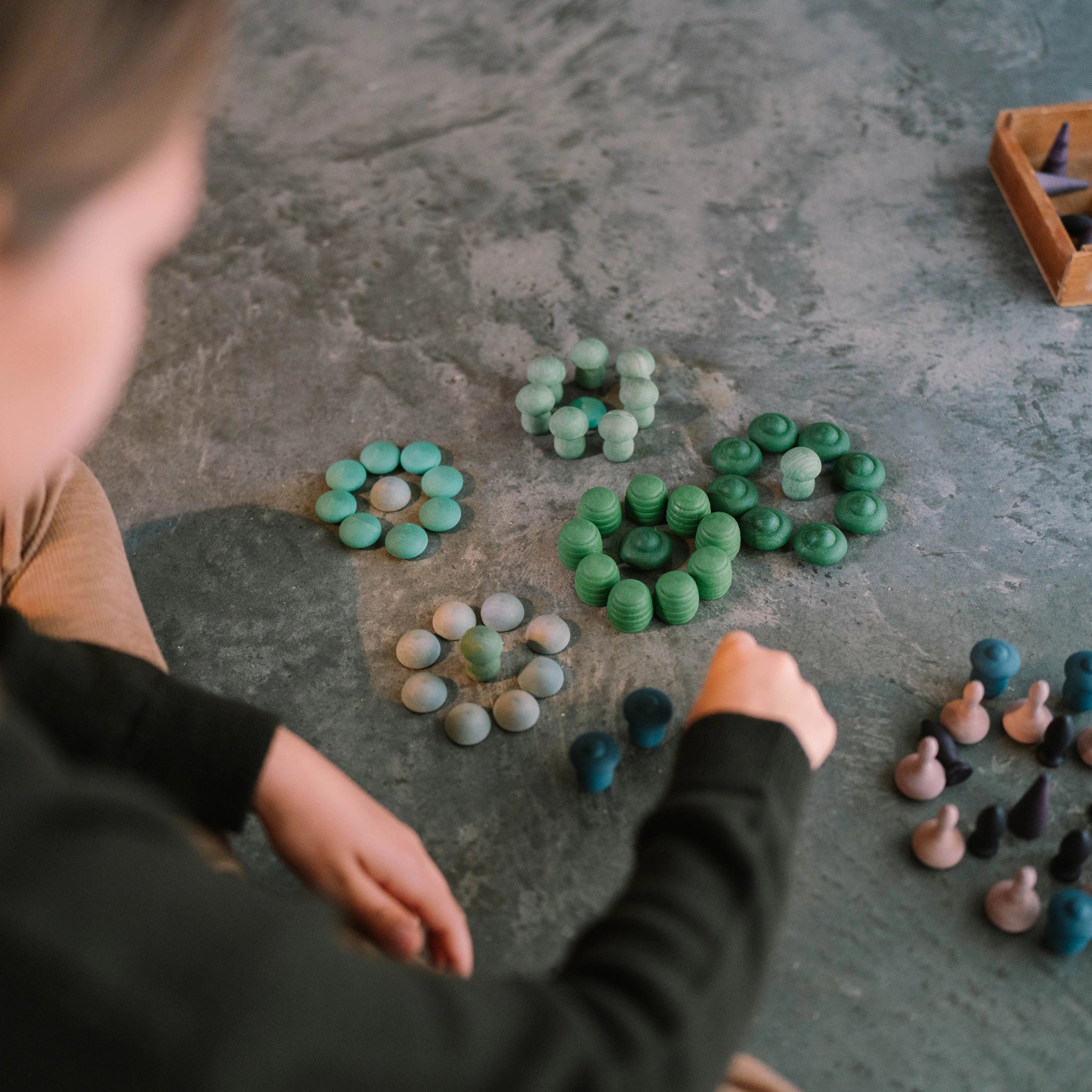 Ein Kind arrangiert beim Freies Spiel: Moonlight zahlreiche, pilzförmige Holzstücke in konzentrischen Kreisen auf einem grauen Untergrund, mit einem kleinen Holztablett voller weiterer Teile im Hintergrund.