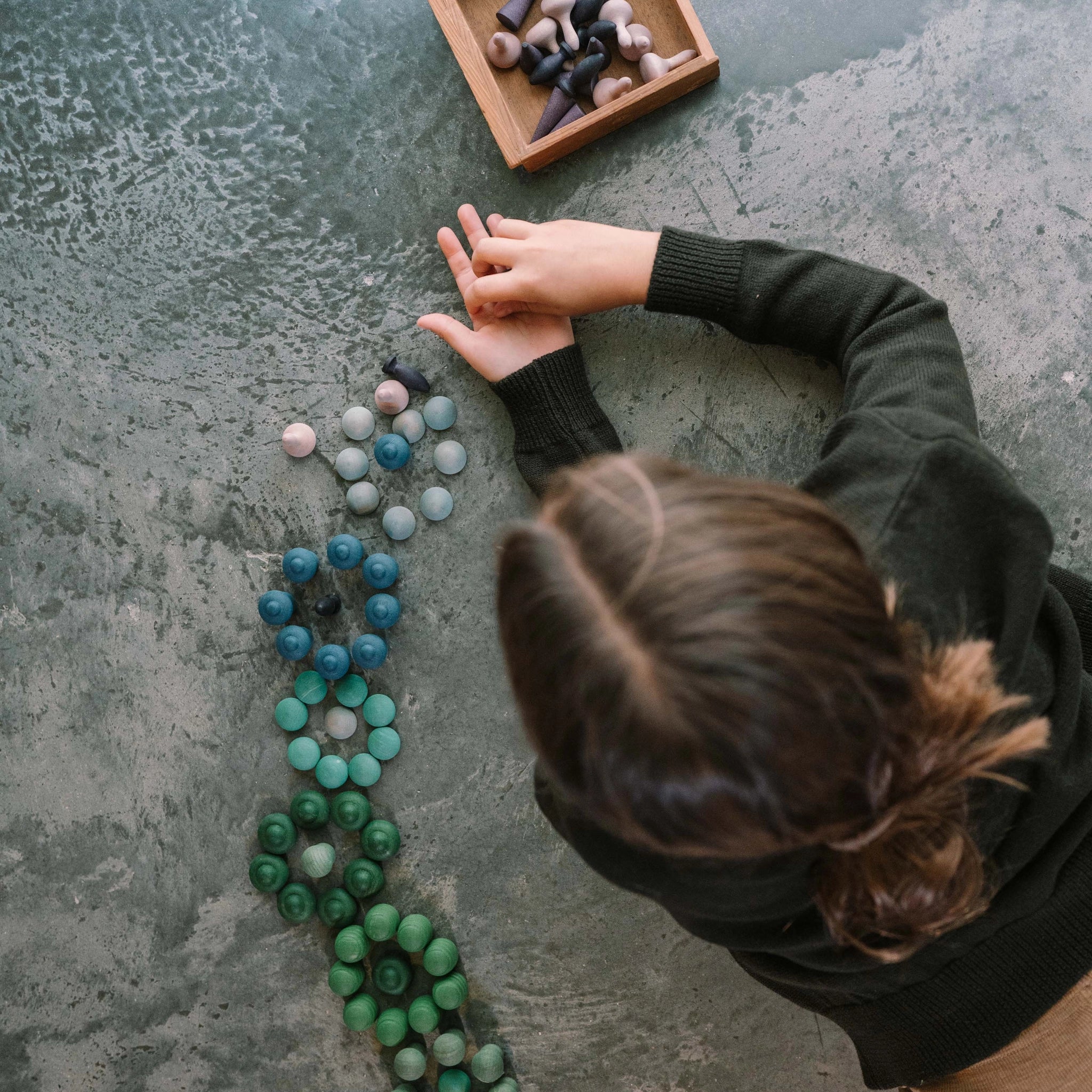 Ein Kind sitzt an einem strukturierten, grauen Tisch und ordnet Freies Spiel: Moonlight Holzspielsteine in Reihen. In einem flachen Holzkasten befinden sich weitere runde Formen, einige pilz- oder kegelartig.