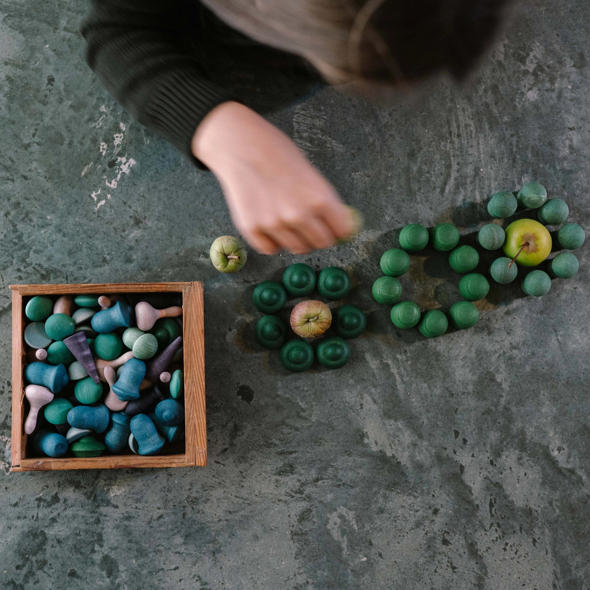 Freies Spiel: Moonlight – Oberansicht zeigt Hände, die hölzerne Teile um Äpfel arrangieren. Links eine offene Box mit weiteren Teilen. Eine Darstellung künstlerischer, spielerischer Holzkomposition von KLICKKLACK.