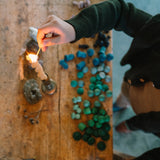 Jemand zündet eine Freies Spiel: Moonlight, verdrehte Tafelkerze im schlichten Metallkerzenhalter an. Sie steht auf einem rustikalen Holztisch, umgeben von blauen und grünen Wachsstücken und natürlichen Elementen.