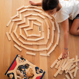 Ein Kind spielt mit Kapla-Bausteine: 200er Box auf hellem Holzboden, arrangiert die gleichförmigen Plättchen in einer spiralförmigen Anordnung. Eine Kapla-Box liegt links unten mit weiteren Bausteinen bereit.