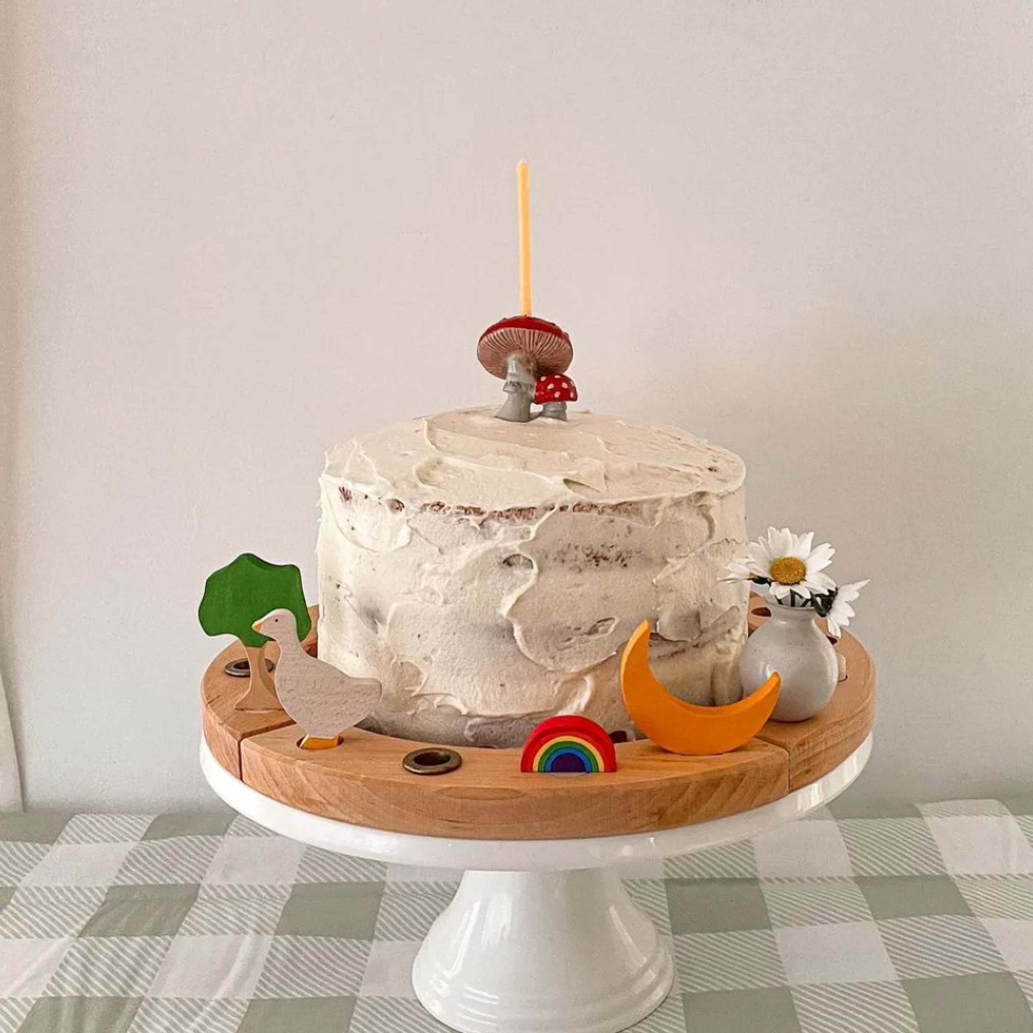 Ein zweischichtiger Naked Cake mit rustikaler Buttercreme wird auf einem weißen Tortenständer präsentiert, dekoriert mit einer Steckfigur: Regenbogen aus handbemaltem Lindenholz, umgeben von weiteren Holzspielzeugen.