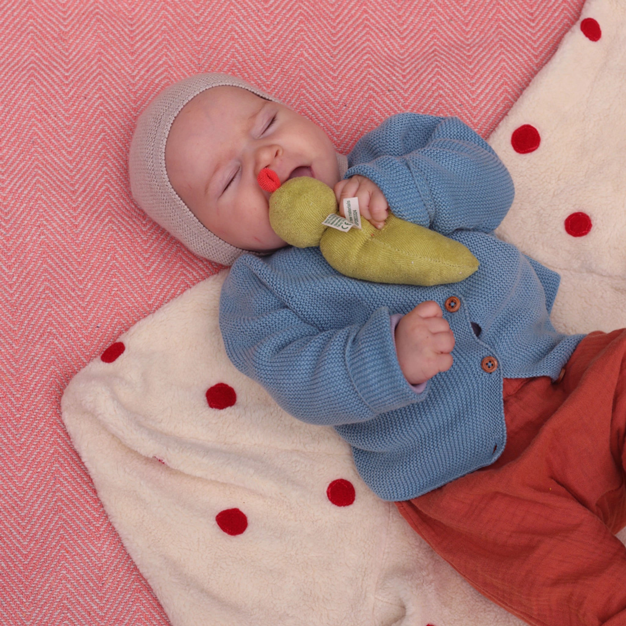 Ein schlafendes Baby hält den Greifling: Bohni, ein grünes Plüschspielzeug mit kleinem rotem Schnabel, während es auf einer weichen Decke liegt, gekleidet in eine gestrickte graue Mütze und blaue Jacke.
