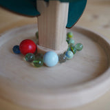 Klangbaum-Spielset: Ein runder Holzteller mit einem vertikalen Stamm und grünen Blättern aus Holz, umgeben von Murmeln, die beim Herabrollen harmonische Klänge erzeugen. Handgefertigt in Österreich.