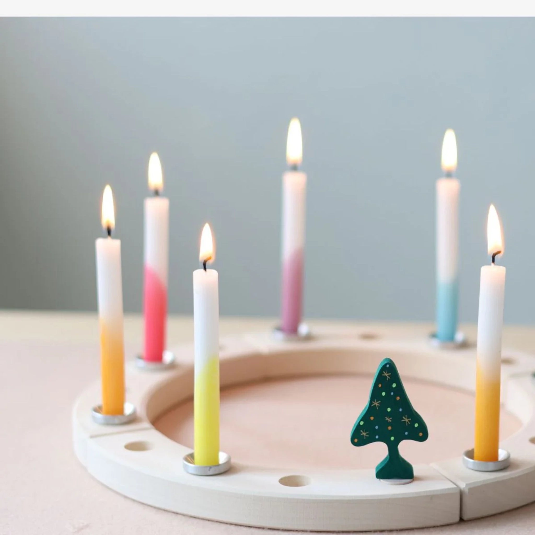 Steckfigur: Dip-Dye Regenbogen Festtags-Kerzen auf einem modularen Holzring mit sieben brennenden Pastellkerzen und dekorativem Baum, ideal für festliche Tischdekorationen, hergestellt in Deutschland aus Bienenwachs und Paraffin.
