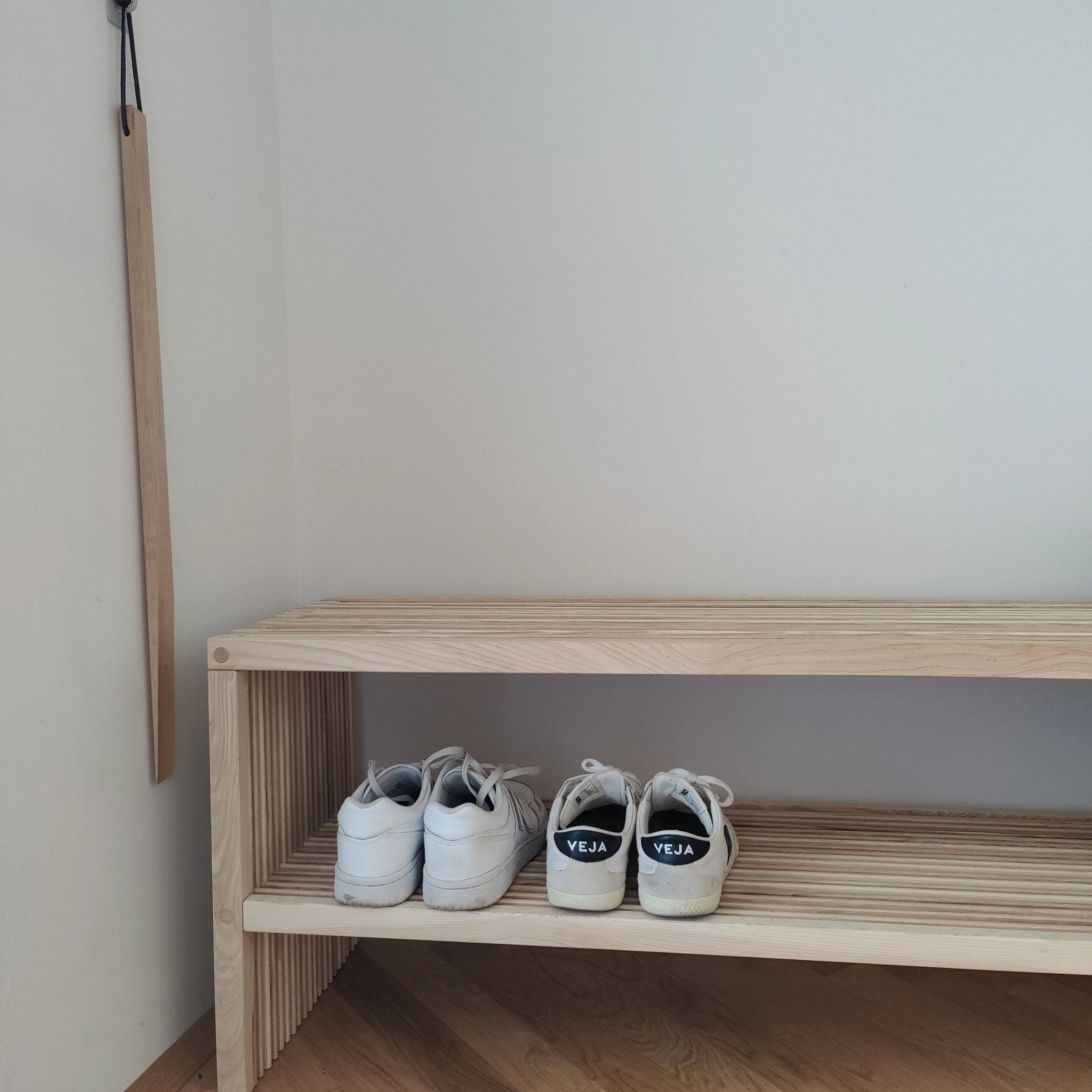 Großer Schuhlöffel aus robustem Buchenholz, hängt an der Wand neben einem leichten Holzregal. Der Schuhlöffel mit Lederband bietet optimale Länge für bequemes Anziehen, handgefertigt in einer geschützten Werkstätte in der Schweiz.