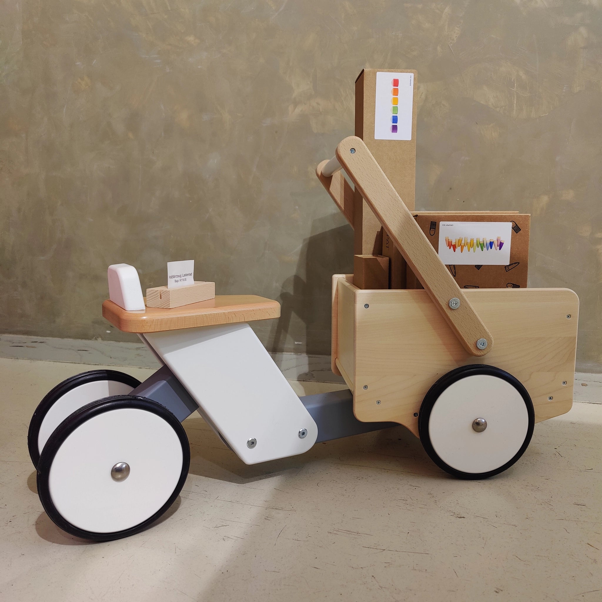 Holzfahrzeug: Lastenrad aus Buchen- und Ahornholz, mit zwei kleinen vorderen und zwei größeren hinteren Rädern, einem Sitz und einer offenen Holzkiste für den Transport von Schätzen, auf glattem Boden.