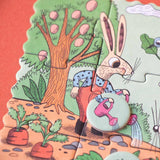 Minipuzzle: Garden zeigt einen Hasen in Latzhose, der mit einer rosa Gießkanne einen kleinen Garten mit Karotten und einem Obstbaum im Hintergrund gießt. Perfekt für Kinder ab 4 Jahren.
