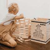 Ein Kind baut mit Bausteine: 280er Kasten von KAPLA einen Turm aus hellen Holzplättchen. Rechts steht eine Kiste mit KAPLA-Plättchen, die Kreativität und logisches Denken fördern.