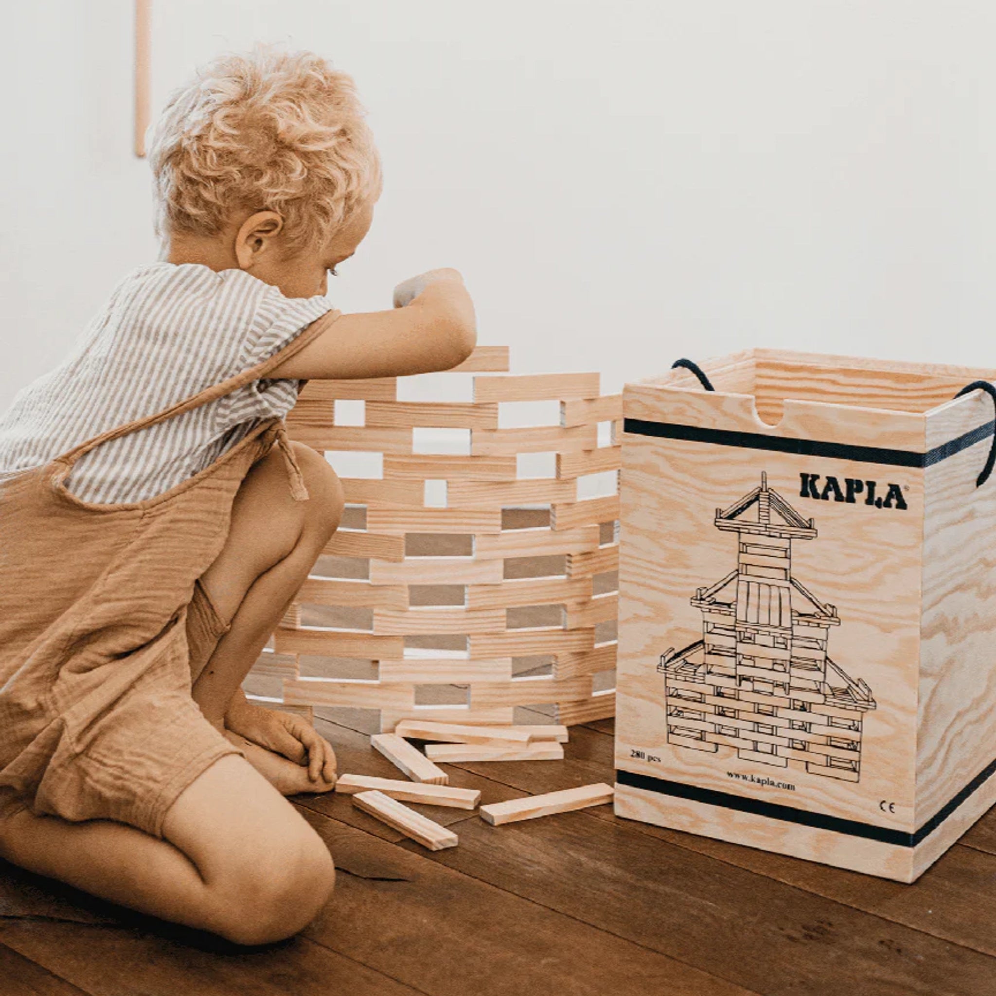 Ein Kind baut mit Bausteine: 280er Kasten von KAPLA einen Turm aus hellen Holzplättchen. Rechts steht eine Kiste mit KAPLA-Plättchen, die Kreativität und logisches Denken fördern.