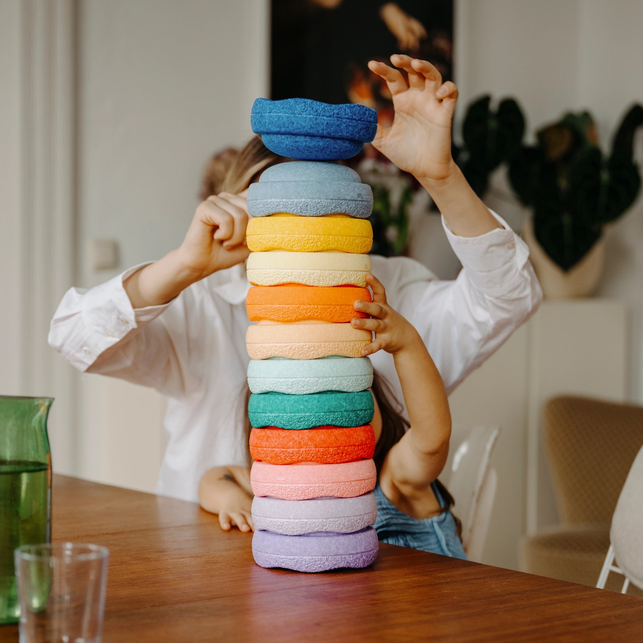 Ein Kind stapelt am Tisch einen hohen Turm aus weichen, strukturierten Ringen des Stapelstein: Mini Regenbogen Classic (6 Steine), während eine erwachsene Hand assistiert.