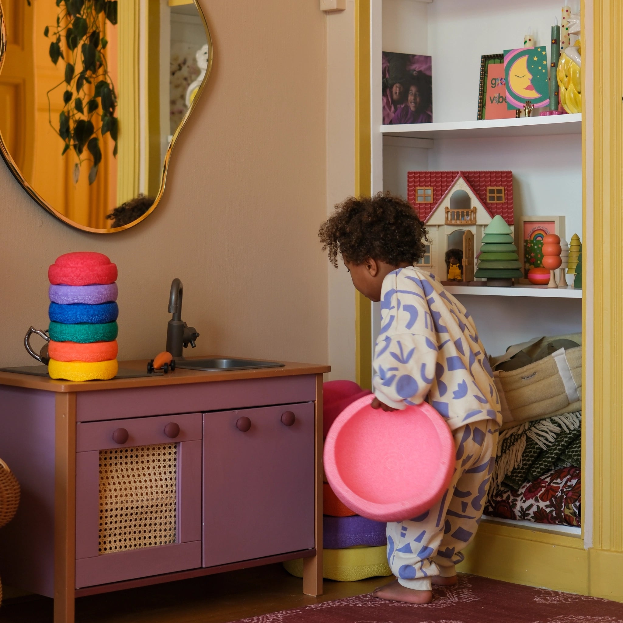 Kind hält einen Stapelstein: Mini Regenbogen Classic (6 Steine) in der Spielküche, arrangiert die bunten Ringe. Im Hintergrund eine Puppenhauslandschaft mit Holzfiguren und Kunstwerken, Spielzimmeratmosphäre von KLICKKLACK.