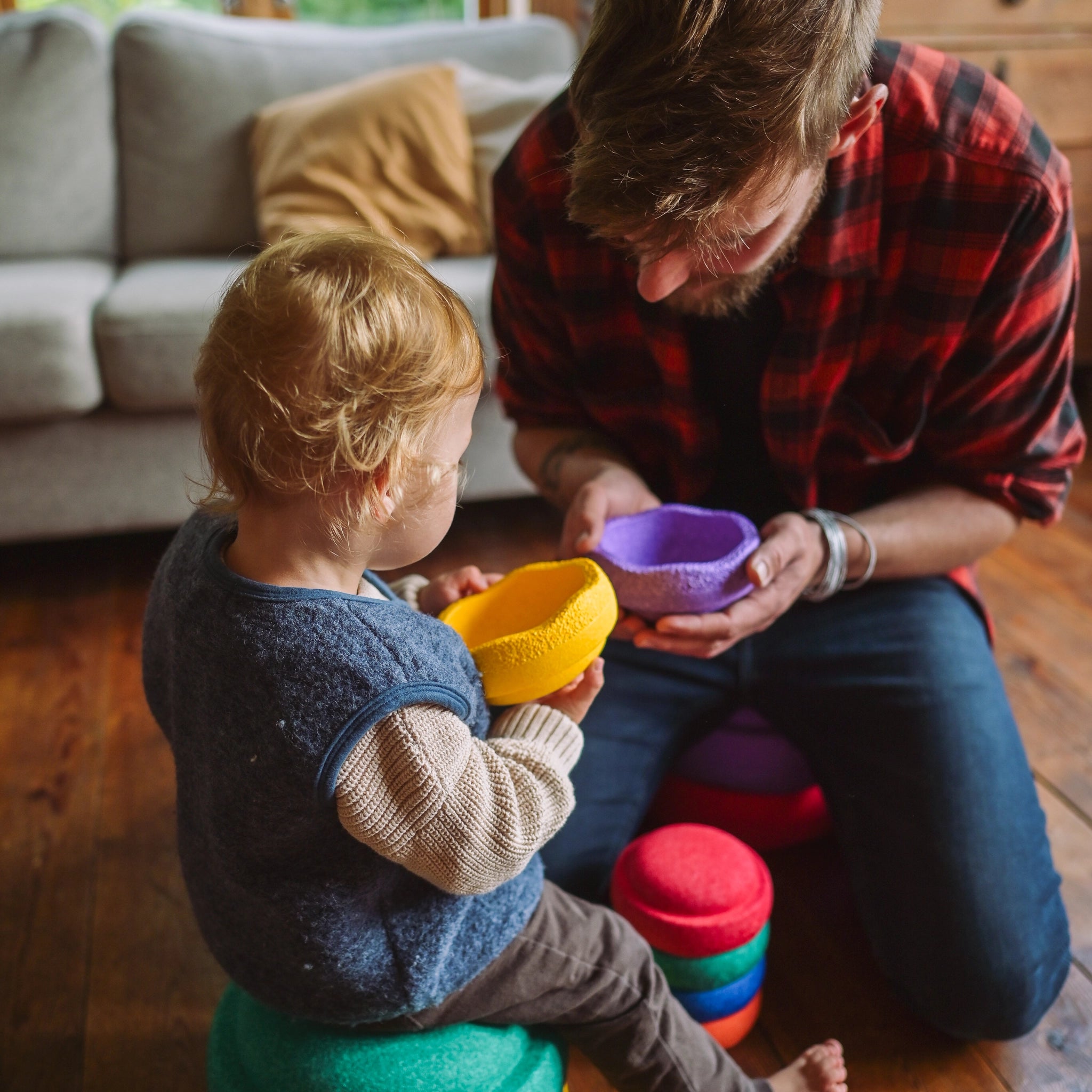 Ein Erwachsener und ein Kind spielen auf dem Wohnzimmerboden mit Stapelstein: Mini Regenbogen Classic (6 Steine), während der Erwachsene ein violettes Teil reicht und das Kind auf einem grünen sitzt.