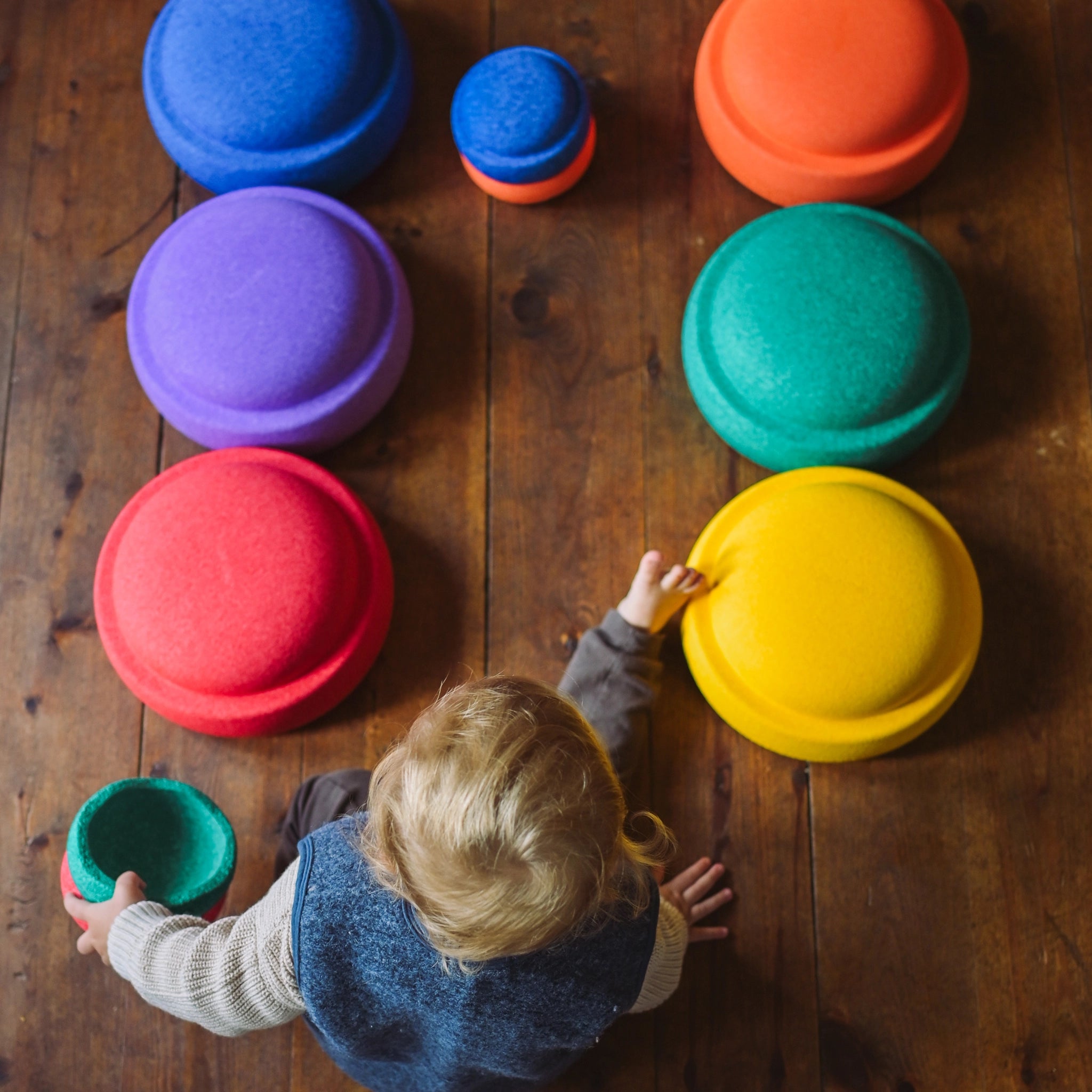Ein Kind spielt auf dem Holzboden mit dem Stapelstein: Mini Regenbogen Classic (6 Steine), indem es einen kleinen grün-roten Becher hält und eine größere gelbe Kuppel berührt.