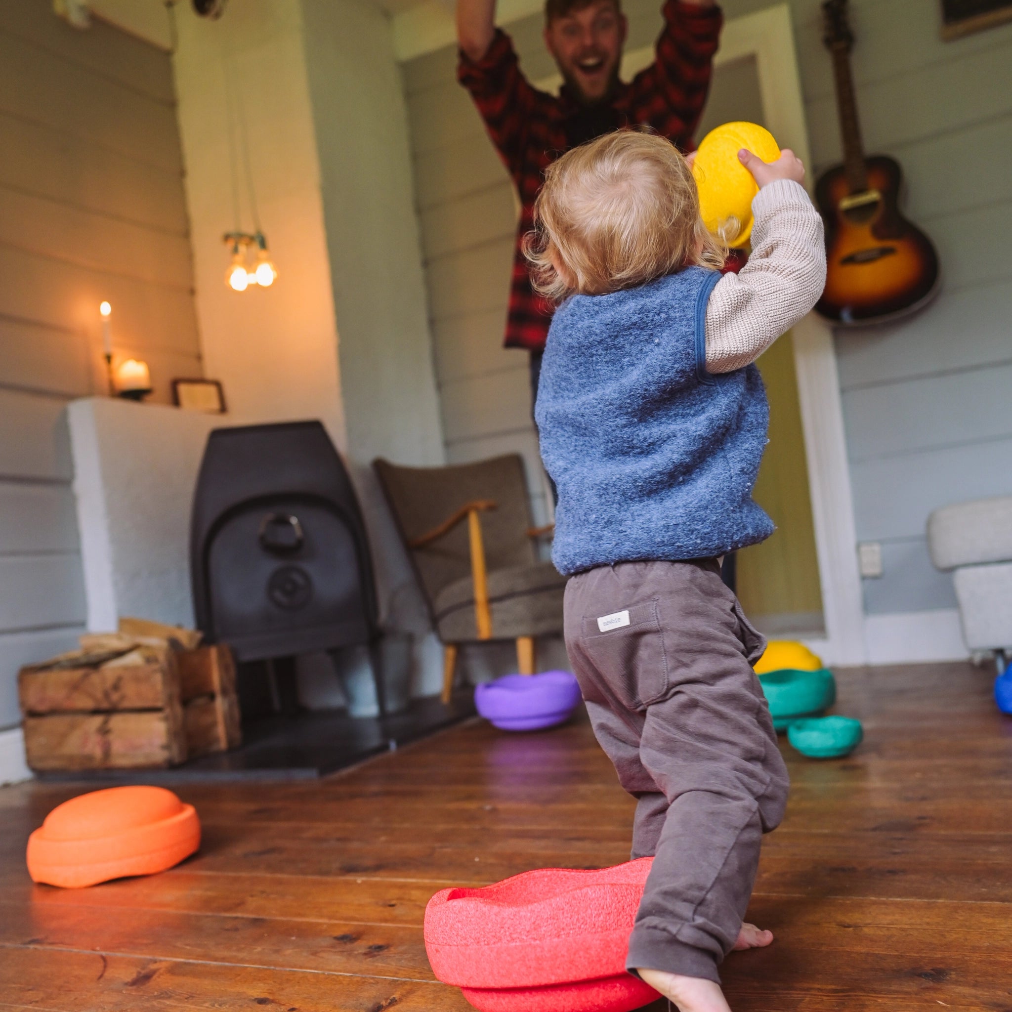 Ein Kleinkind balanciert auf einem Stapelstein: Mini Regenbogen Classic (6 Steine), gelber Schaumstoff, auf einem Wohnzimmerboden. Im Hintergrund jubelt ein Erwachsener, während weitere farbige Steine auf dem Boden verteilt sind.