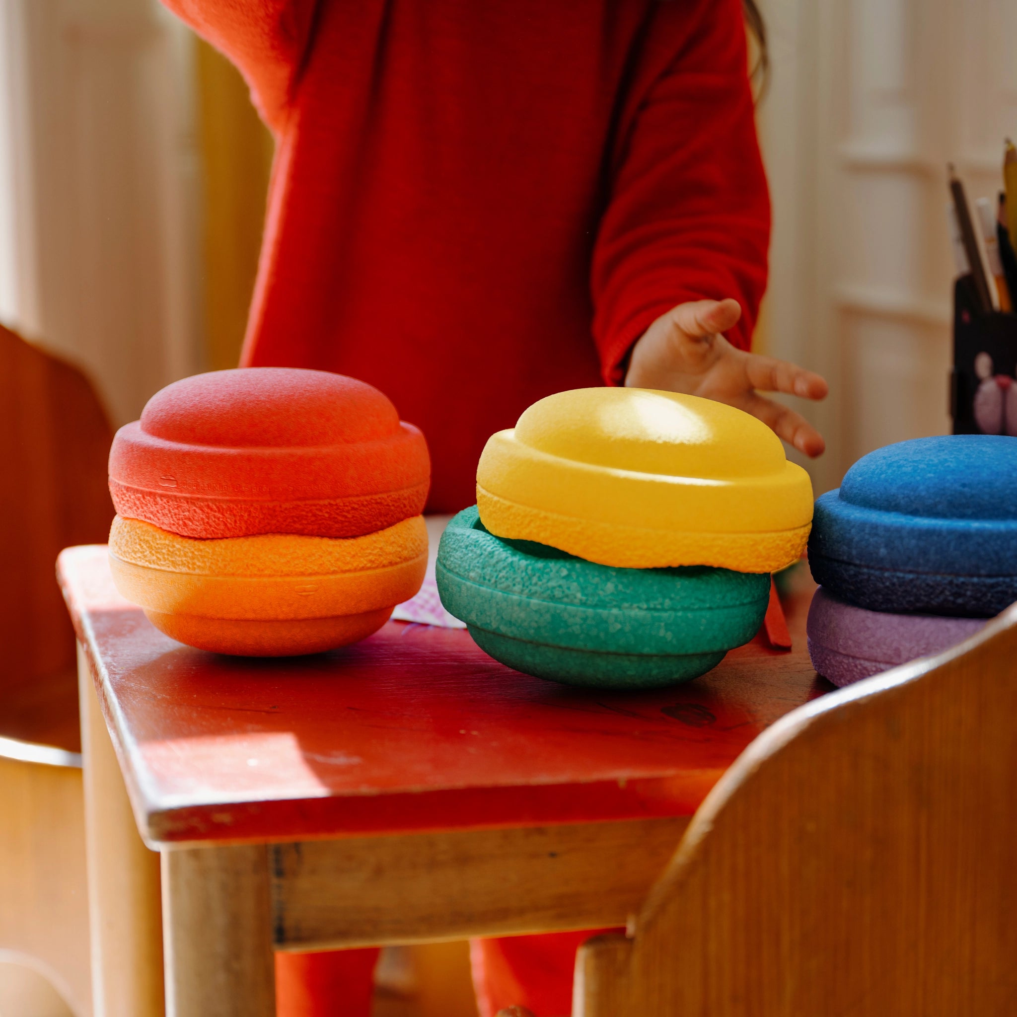 Stapelstein: Mini Regenbogen Classic (6 Steine) auf einem kleinen Tisch im Spielbereich, mit einem Kind, das nach einem der bunten, stapelbaren Schaumstoffspielzeuge greift.