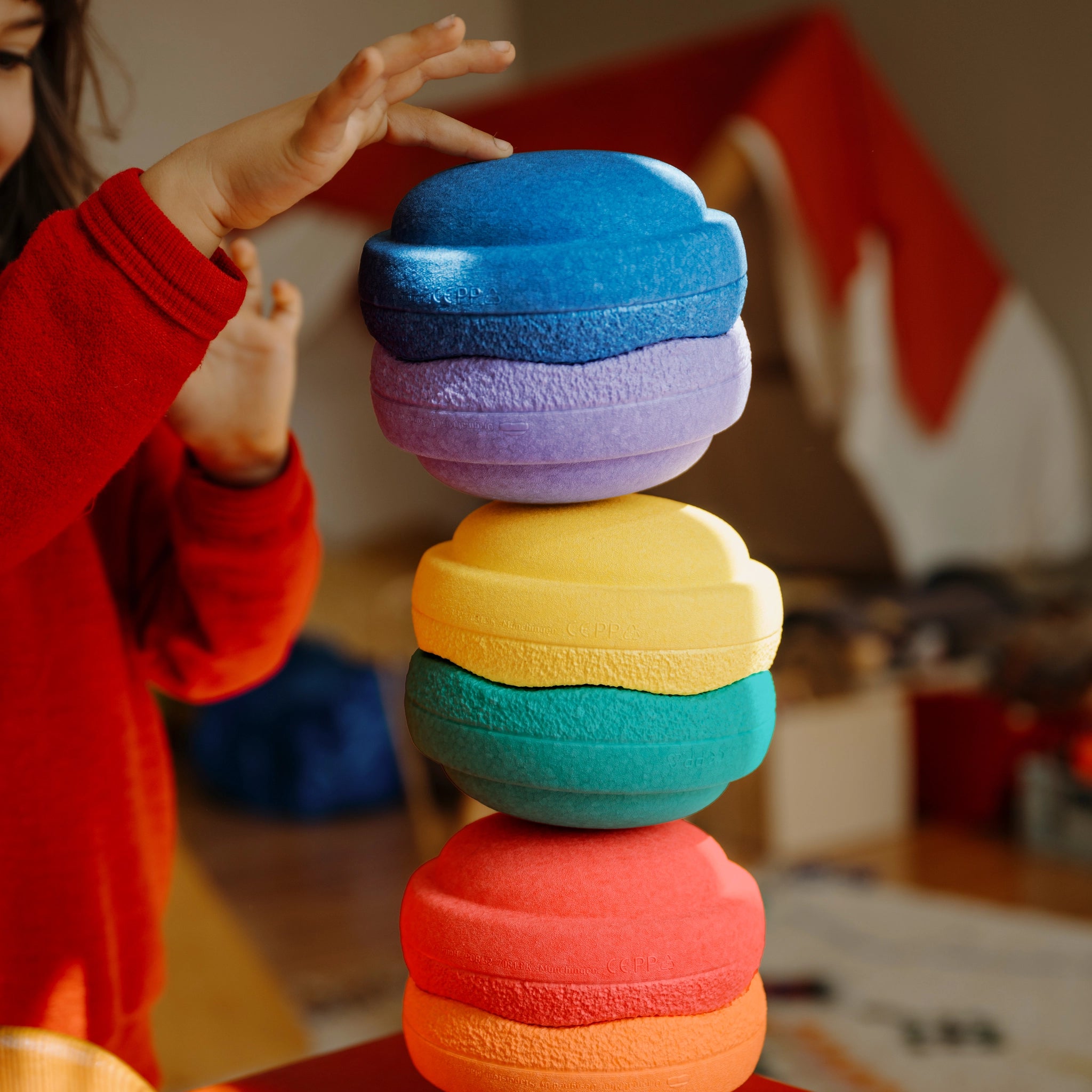 Kind zeigt, wie es sechs bunte Schaumstoffsteine des Stapelstein: Mini Regenbogen Classic (6 Steine) balanciert. Die Steine sind nachhaltig und fördern kreatives Spielen im Indoor-Bereich.