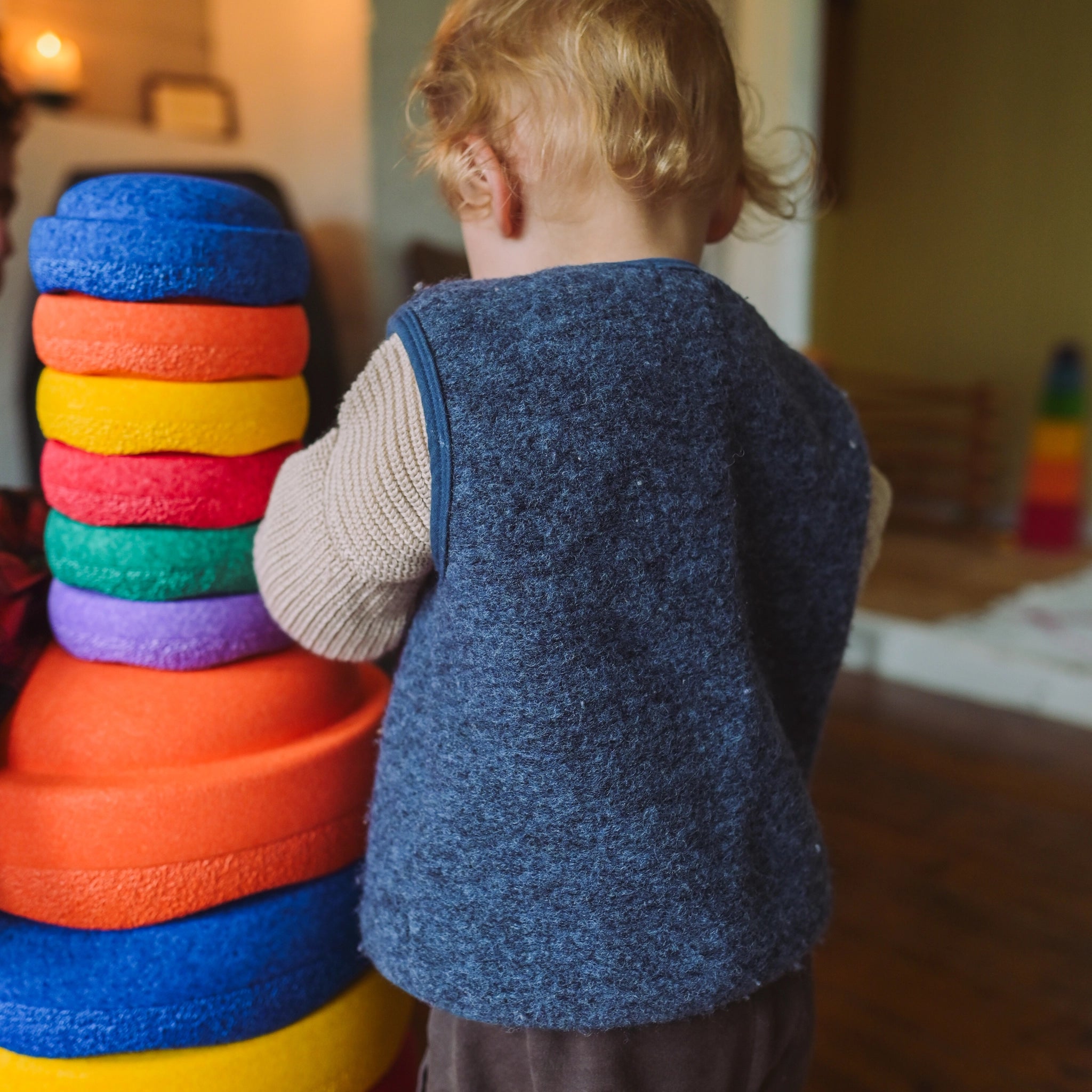 Ein Kleinkind stapelt von hinten gesehen eine Säule aus großen, bunten Schaumringen. Der Stapelstein: Mini Regenbogen Classic (6 Steine) zeigt lebendige Farben im Spielbereich, ideal für kreatives Spiel.