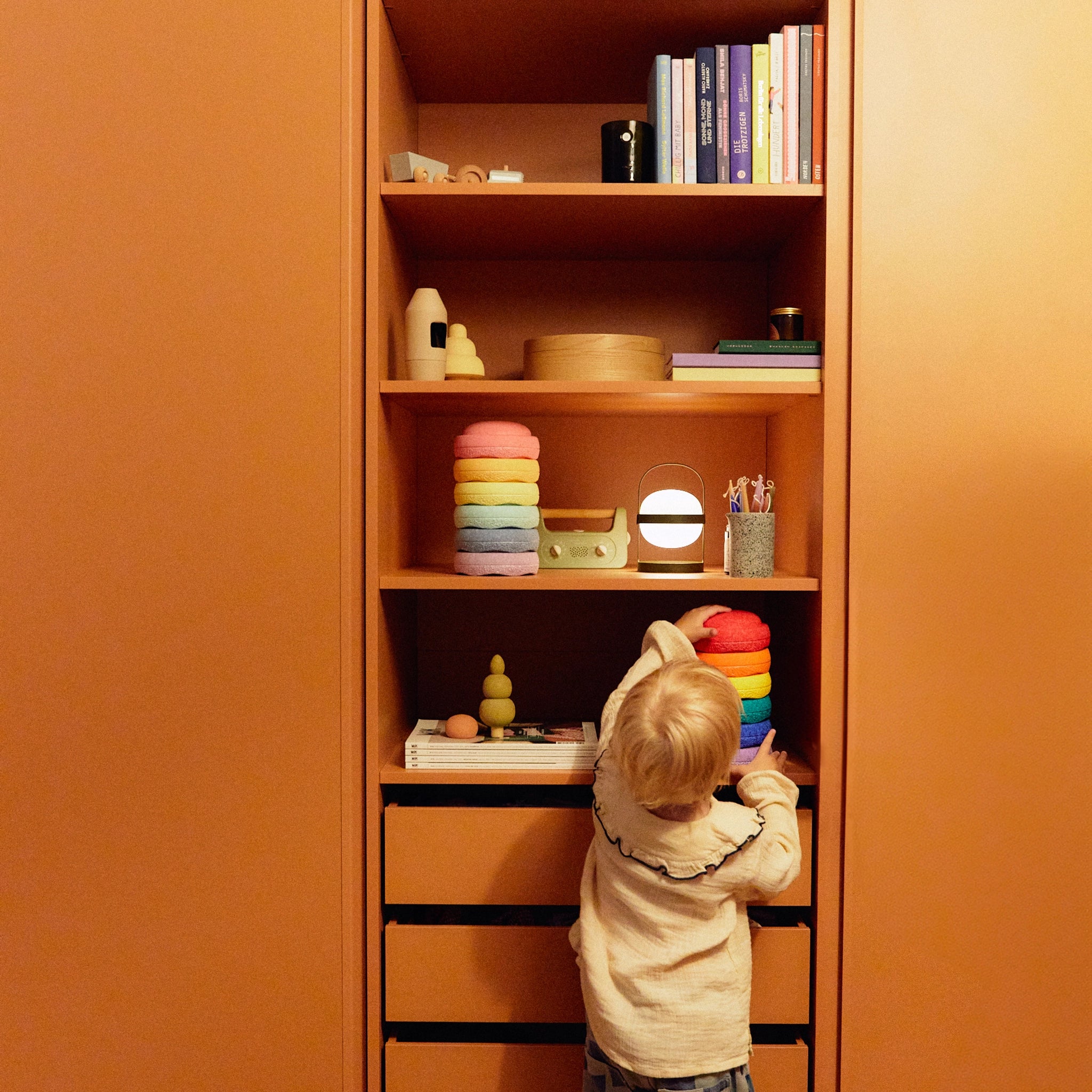 Ein Kleinkind greift nach einem Stapelstein: Mini Regenbogen Classic (6 Steine) in einem offenen Regal, umgeben von modernen Spielzeugen und Büchern, passend zur ästhetischen und nachhaltigen Ausrichtung von KLICKKLACK.
