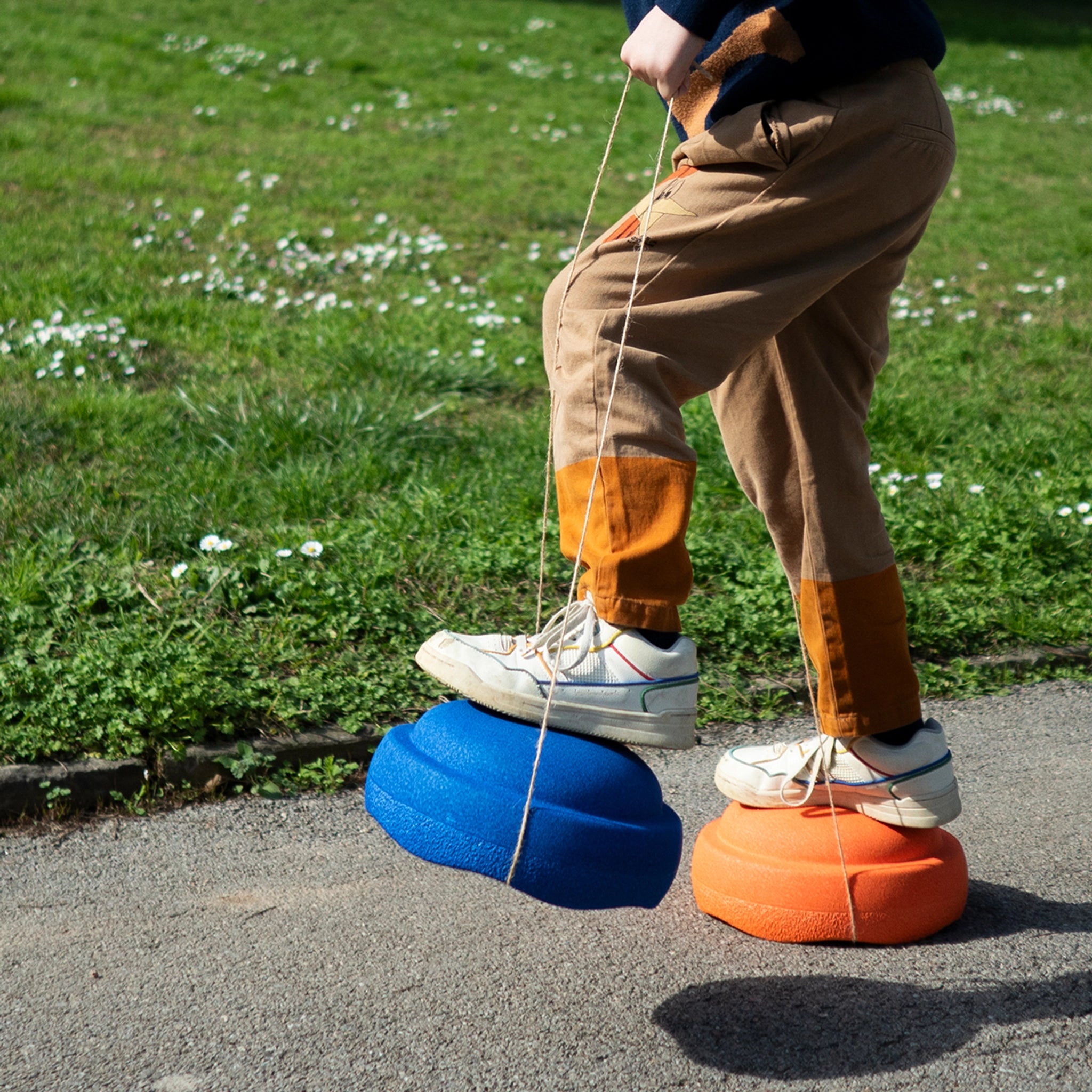 Ein Kind balanciert auf Stapelstein: Regenbogen Classic (6 Steine) aus Kunststoff, befestigt an den Füßen mit Schnüren, während es auf einem gepflasterten Weg neben einer Wiese mit weißen Blumen geht.