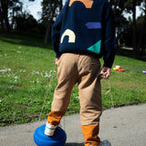 Ein Kind balanciert auf zwei Stapelstein: Orange, runden blauen Trittsteinen im Park, hält Seile in den Händen. Es trägt helle Sneaker, tanfarbene Hosen und einen dunklen Pullover mit abstrakten Formen.