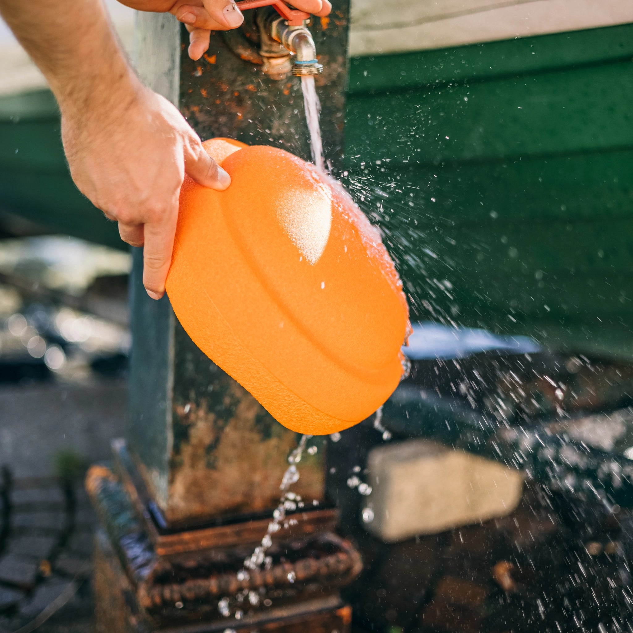 Eine Person hält einen orangefarbenen Teller aus der Stapelstein: Regenbogen Classic (6 Steine) Kollektion unter einem Außenwasserhahn, während Wasser darüber fließt und gegen einen verwitterten Metallpfosten spritzt.