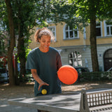 Eine junge Person spielt fröhlich an einem Outdoor-Tischtennistisch, hält einen Stapelstein: Orange in der Hand und jongliert einen gelben Ball. Im Hintergrund sind Bäume und ein Gebäude zu sehen.