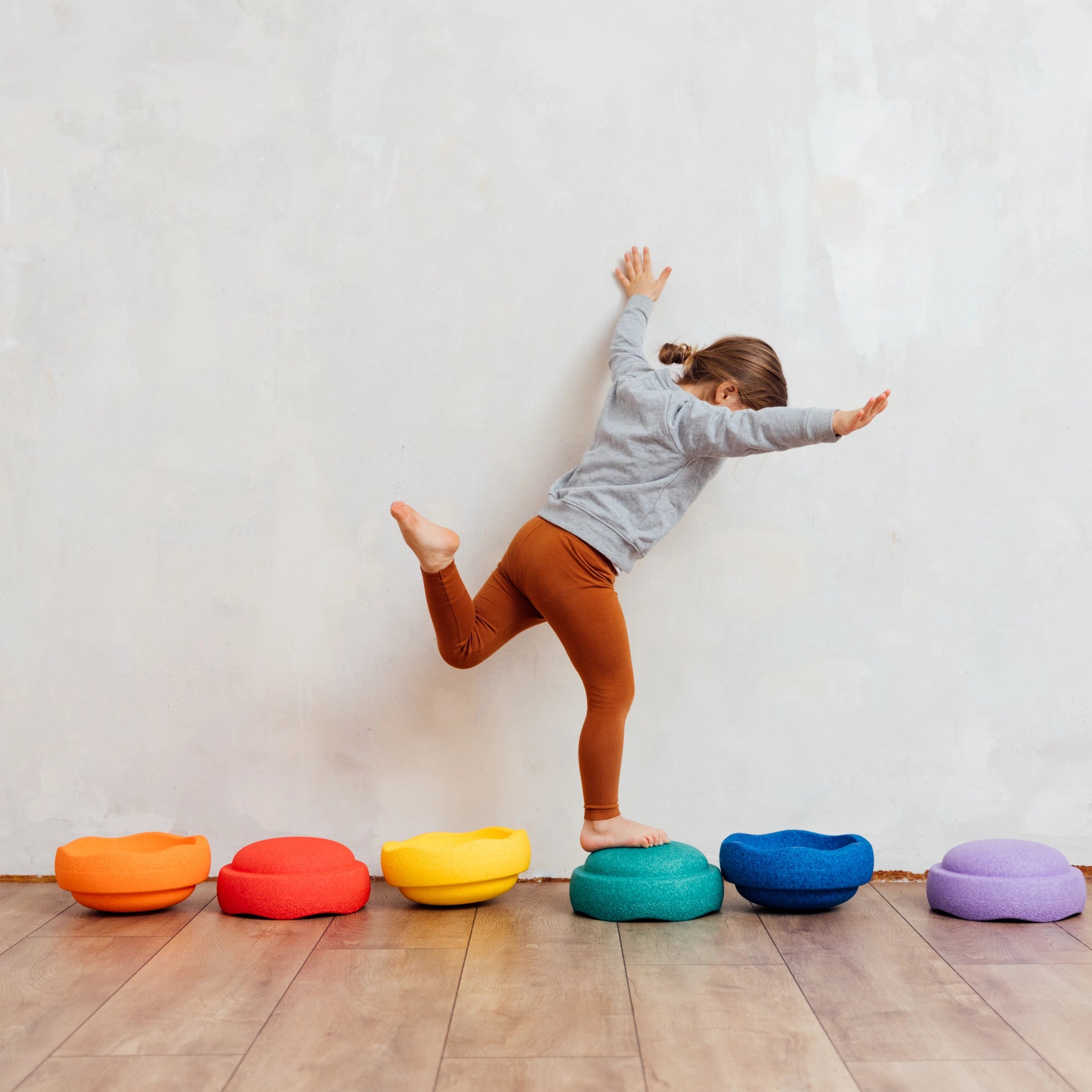 Ein Kind balanciert spielerisch auf einem grünen Stapelstein: Regenbogen Classic (6+1 Balance Board), einer Reihe bunter Schaumstoffkissen, die Bewegung und Gleichgewichtssinn fördern und nachhaltige Freude bieten.