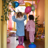 Zwei Kinder mit Partyhüten stehen auf Stapelstein: Regenbogen Classic (6+1 Balance Board) in einem festlich dekorierten Wohnzimmer, umgeben von Luftballons, vermitteln Freude und Nachhaltigkeit für moderne, umweltbewusste Kunden.