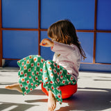 Ein Kind sitzt auf dem Stapelstein: Regenbogen Classic (6 Steine) roten Kissen, trägt ein rosa Shirt und grün-weiß karierte Hose, vor blauen Paneelen, verkörpert nachhaltiges, modernes Design von KLICKKLACK.