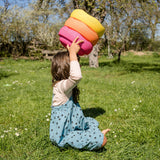 Ein Kind kniet auf dem Rasen und balanciert Stapelstein: Regenbogen Classic (6 Steine) auf dem Kopf. Im Hintergrund sind Bäume und eine Holzbank sichtbar, was auf Spielen im Freien hindeutet.